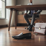 ARQS.PK black textured leather slip-on loafers on a wooden office floor with a businessman in the background.